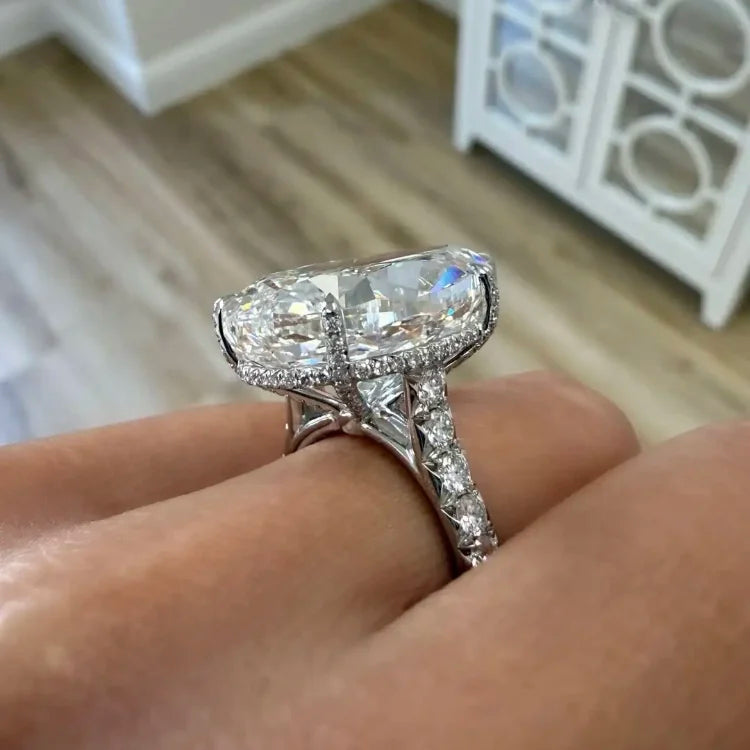 A close-up of a hand wearing a large oval diamond ring with a silver-toned setting and pavé accents, photographed indoors against a blurred background of wood flooring and a white cabinet.