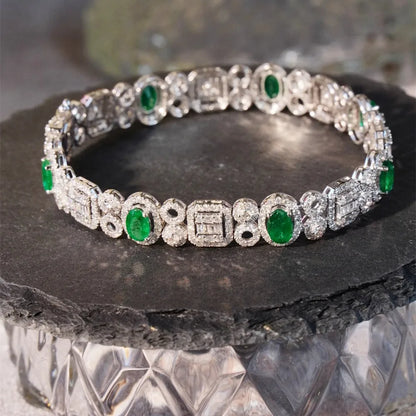 An ornate silver-toned tennis bracelet with round green emerald-cut gemstones and pavé-set clear stones, displayed on a dark stone slab.