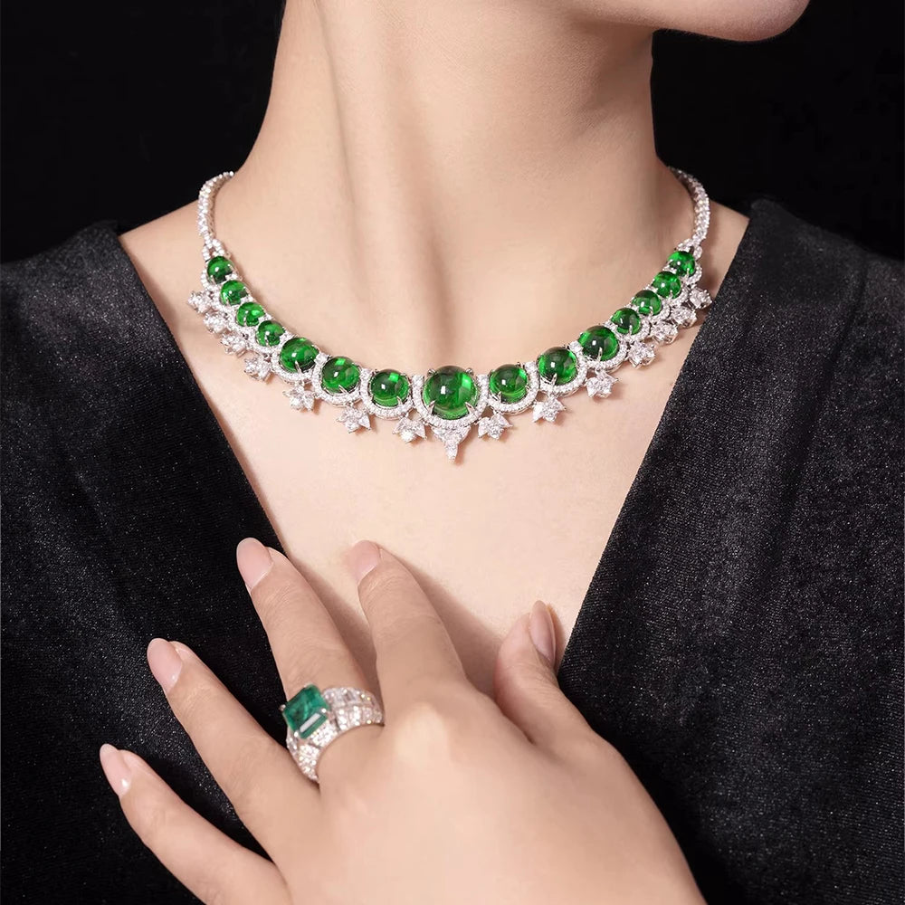 A close-up lifestyle shot of a model wearing a silver-toned necklace with large round green emerald-cut gemstones and a matching emerald ring on her finger, set against a black velvet top and a dark background.