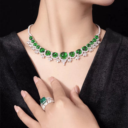 A close-up lifestyle shot of a model wearing a silver-toned necklace with large round green emerald-cut gemstones and a matching emerald ring on her finger, set against a black velvet top and a dark background.
