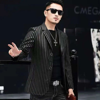 A man wearing a black and gray vertically striped blazer over a black shirt, accessorized with a silver cross necklace, sunglasses, and a decorative belt, posing in a dark setting.