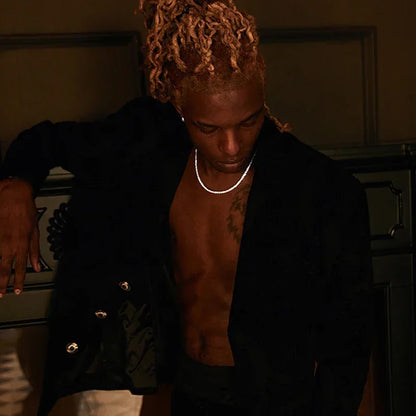 A man with blonde dreadlocks leans against a dark cabinet, wearing an open black blazer over a bare torso, a silver chain necklace, and white pants in a dimly lit interior.