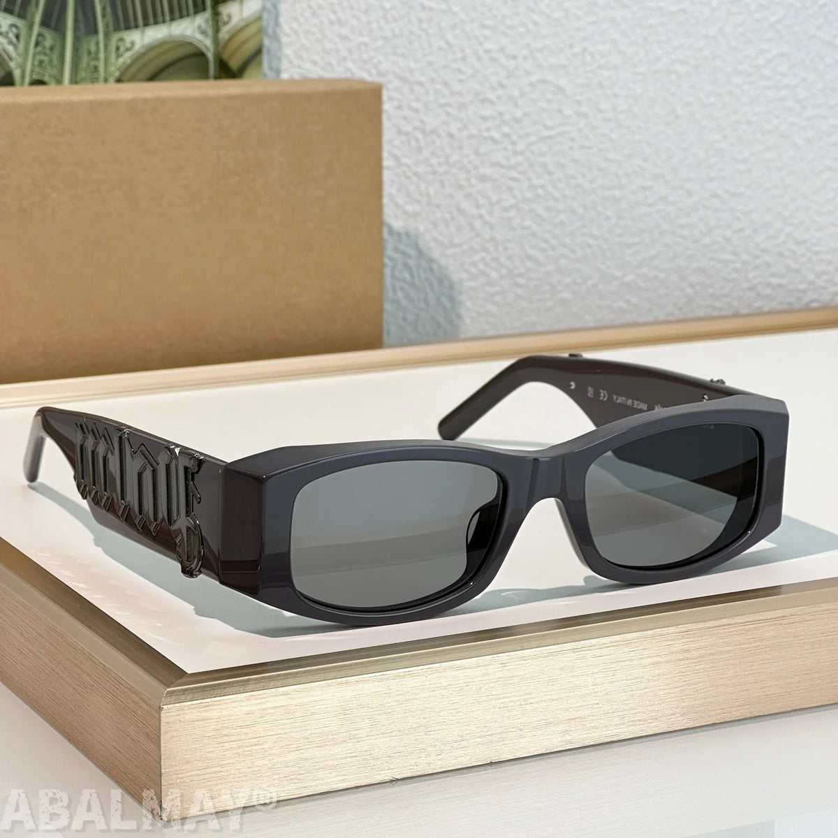 Black square sunglasses with dark lenses and sculptural temple details, displayed on a light wood tray with a brown box in the background.