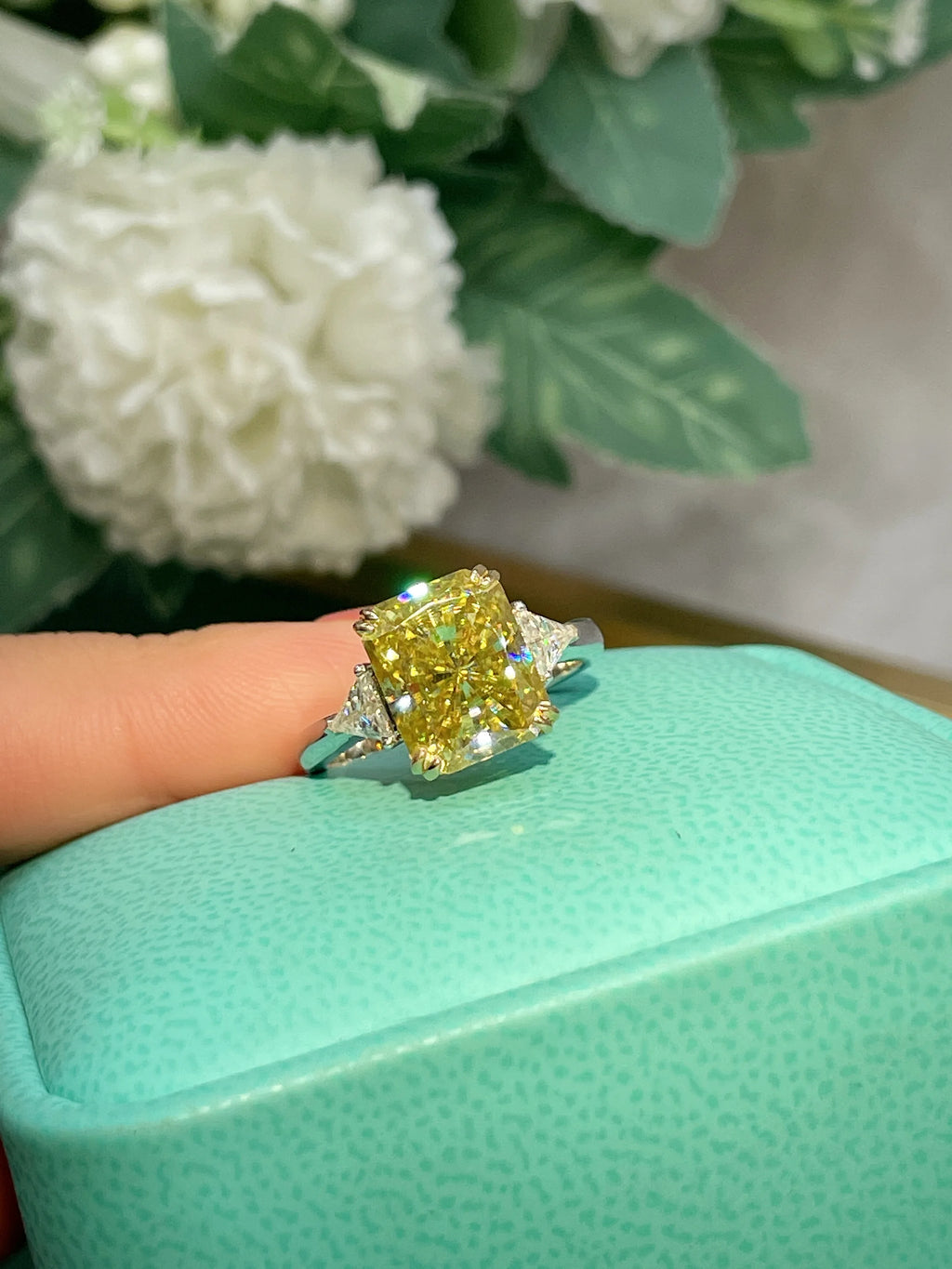 A yellow diamond ring with a cushion-cut center stone and smaller side stones on a silver-toned band, shown resting on a mint green textured box with a finger holding it. White flowers and green leaves are blurred in the background.