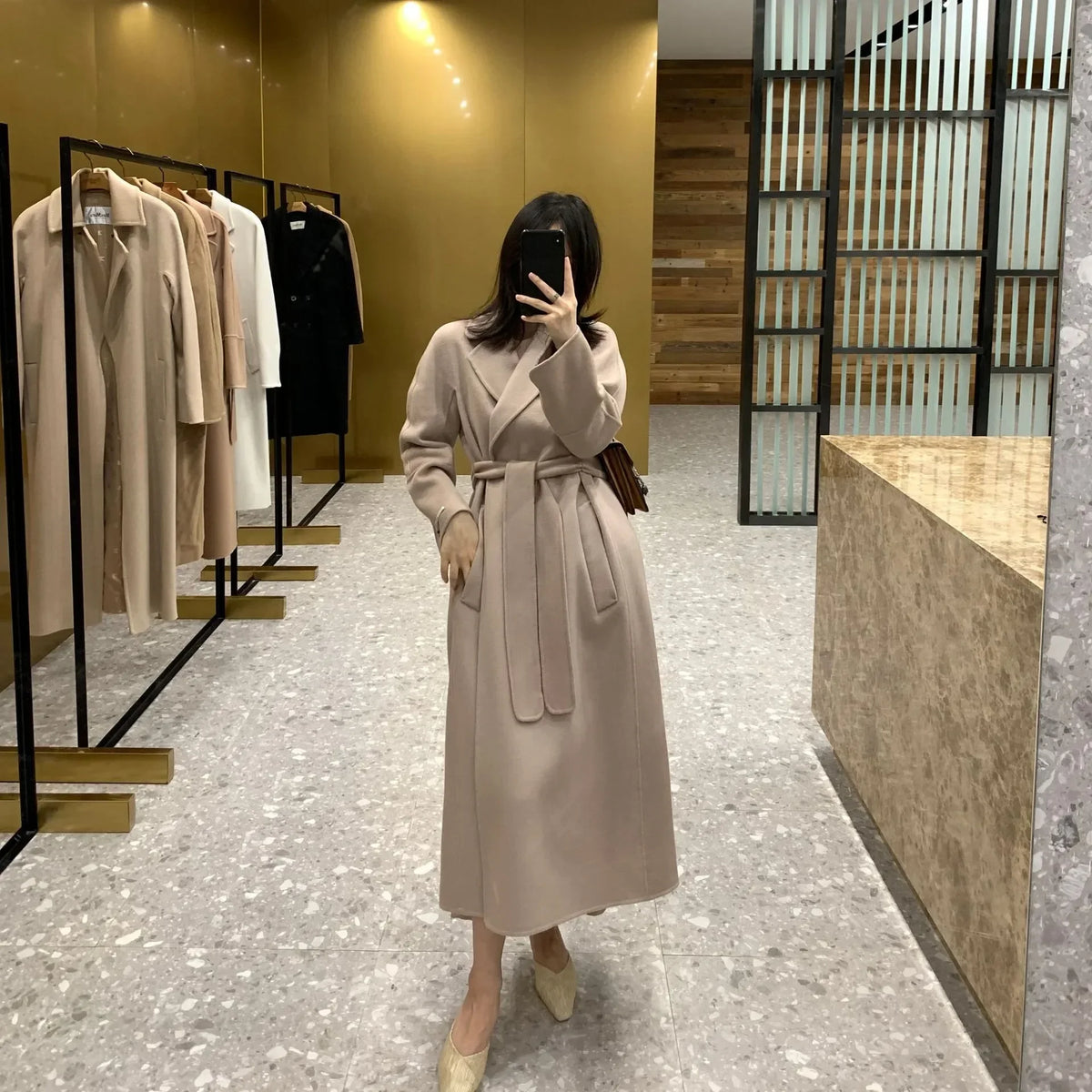 A woman takes a mirror selfie in a boutique wearing a long beige belted coat. The boutique has gold-toned walls, clothing racks with neutral-colored garments, and a marble counter.