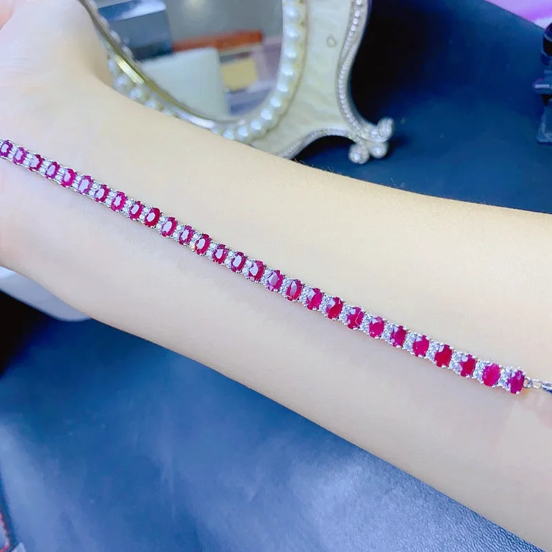 A close-up of a silver-toned tennis bracelet with rectangular red gemstones and small clear accent stones, worn on a person's wrist against a dark blue surface.