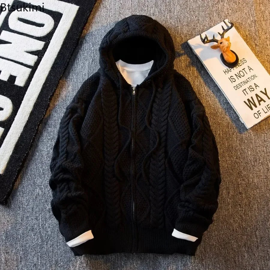 A black cable-knit zip-up hoodie with a hood, laid flat on a gray textured surface. The hoodie features a ribbed hem and cuffs with white lining peaking out at the openings.