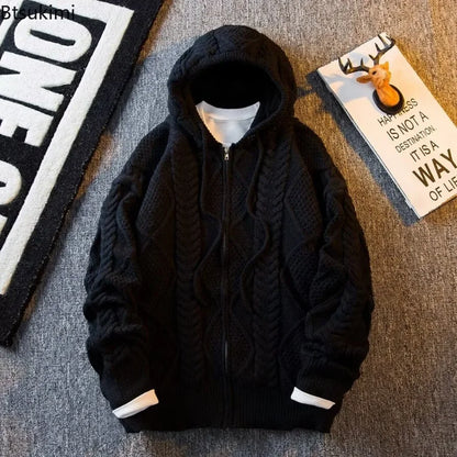 A black cable-knit zip-up hoodie with a hood, laid flat on a gray textured surface. The hoodie features a ribbed hem and cuffs with white lining peaking out at the openings.