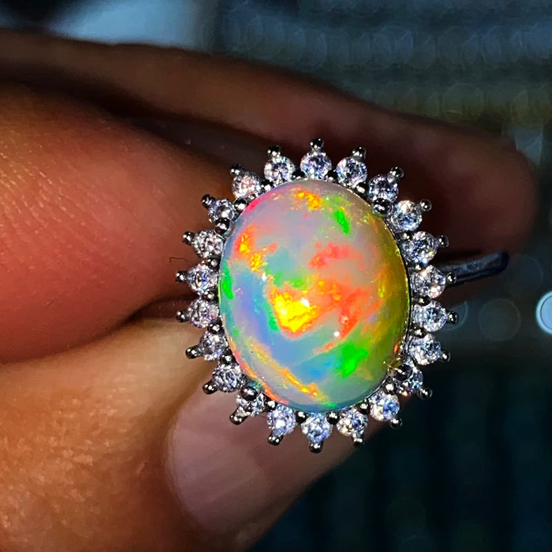 A close-up of a ring being held on fingers, featuring a round iridescent opal center stone surrounded by a halo of small clear stones in a silver-toned setting.