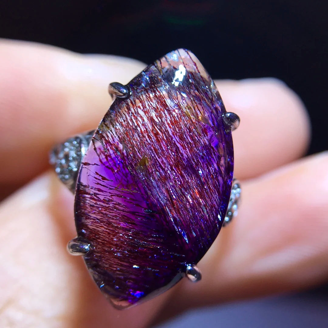 A close-up of a hand holding a silver ring with a large teardrop-shaped purple and iridescent gemstone featuring fine metallic needle-like inclusions.