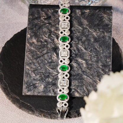 A silver-toned bracelet with alternating oval green emerald-cut stones and square diamond-cut stones, displayed on a dark textured square block.