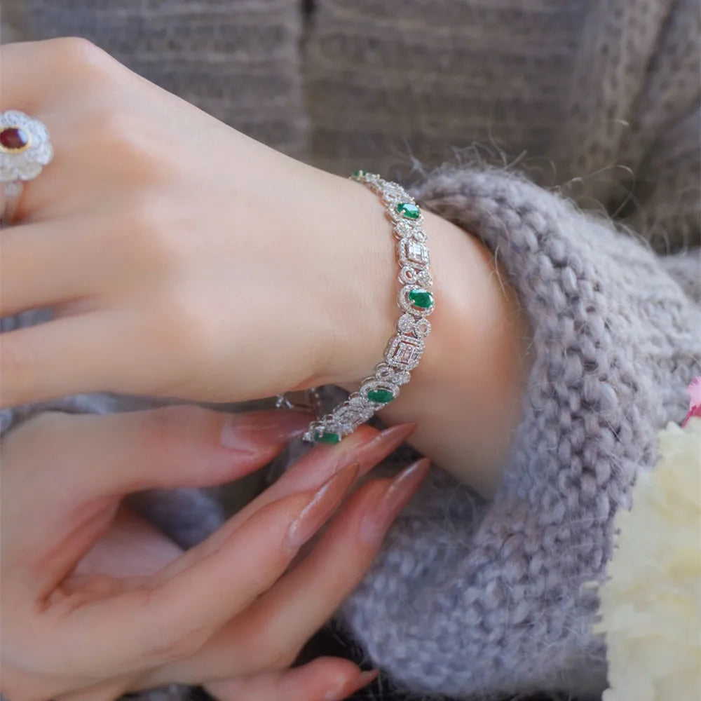 A close-up of a person's wrist wearing a silver-toned bracelet adorned with green emerald-cut stones and clear crystals, paired with a chunky gray knit sweater.