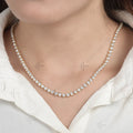 A close-up of a model wearing a delicate gold tennis necklace set with clear round-cut gemstones, styled with a white collared shirt.
