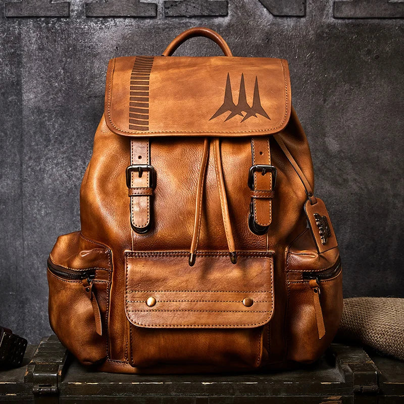 A brown distressed leather backpack with a front flap pocket featuring an engraved mountain-like logo, buckle straps, and side pockets, displayed on a rustic wooden crate against a dark textured wall.