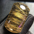 A rectangular clear gemstone with dense golden hair-like inclusions, photographed close up against a dark textured fabric background.