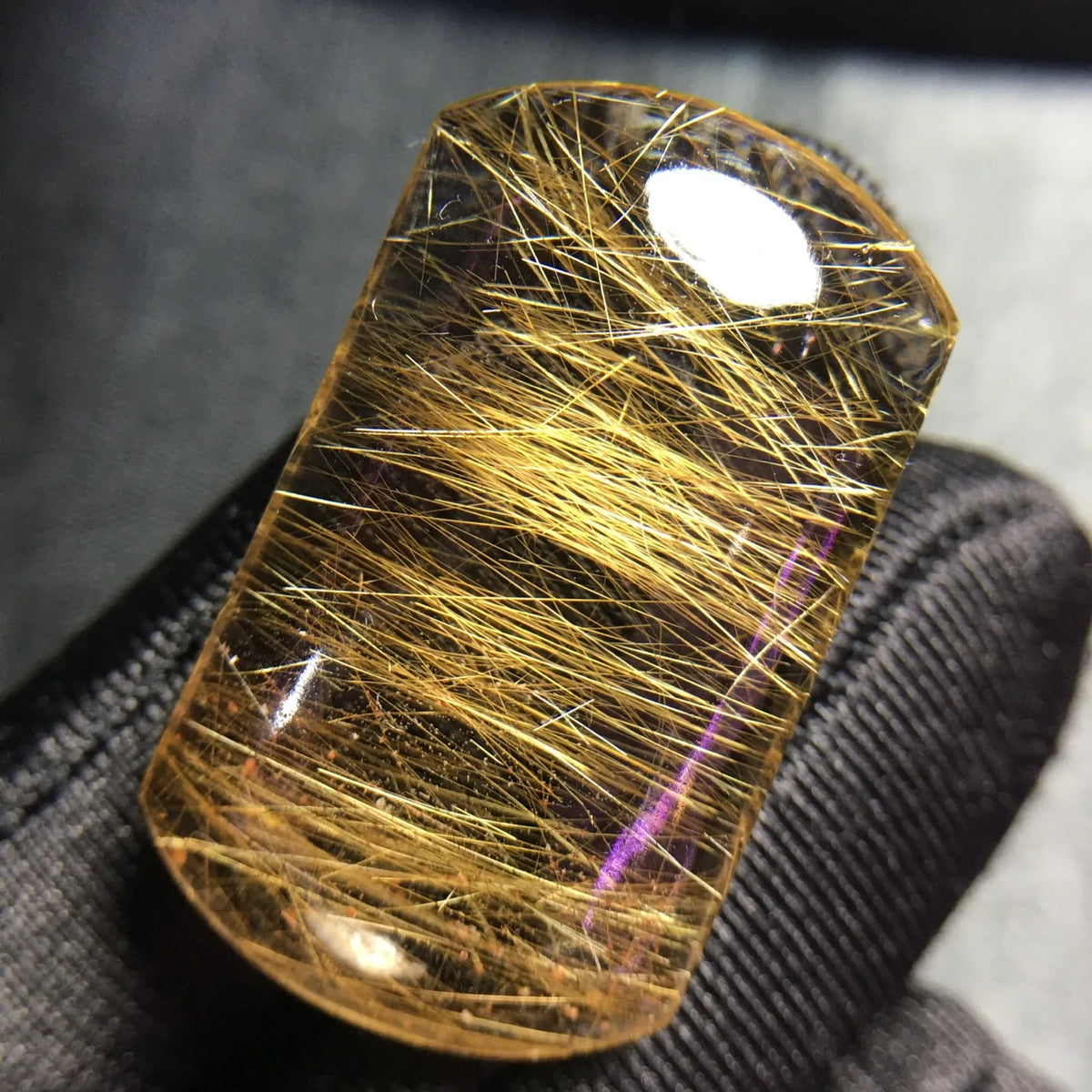 A rectangular clear gemstone with dense golden hair-like inclusions, photographed close up against a dark textured fabric background.