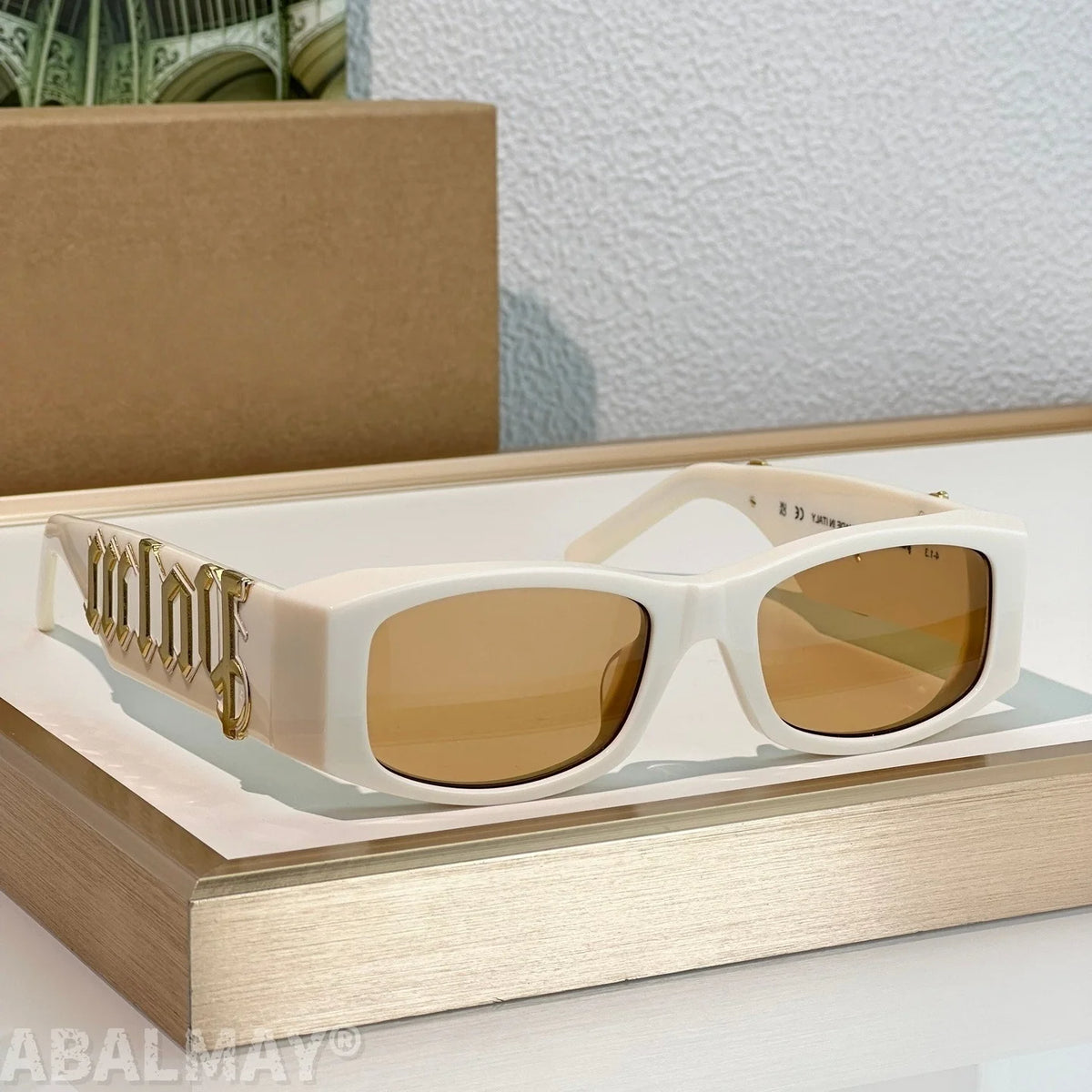 White square sunglasses with gold-tinted lenses and a decorative gold script detail on the temple arm, displayed on a light wood tray.