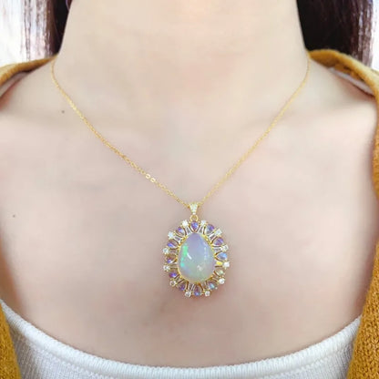 A gold chain necklace with a teardrop-shaped iridescent opal pendant, worn on a model's neck. The pendant is surrounded by small clear stones and lavender-purple accent stones, set against a model's skin with a mustard yellow cardigan visible.