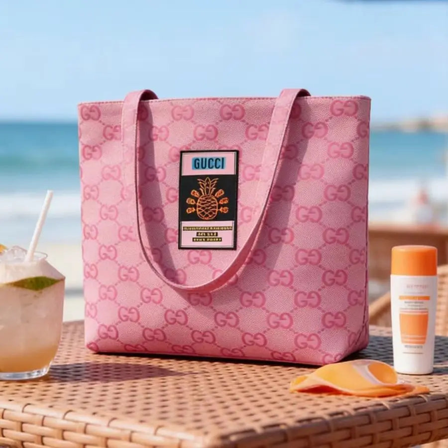A pink Gucci tote bag with an allover GG monogram pattern and a central Gucci patch, displayed on a wicker table at a beach with a drink, towel, and sunscreen nearby.