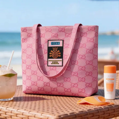 A pink Gucci tote bag with an allover GG monogram pattern and a central Gucci patch, displayed on a wicker table at a beach with a drink, towel, and sunscreen nearby.