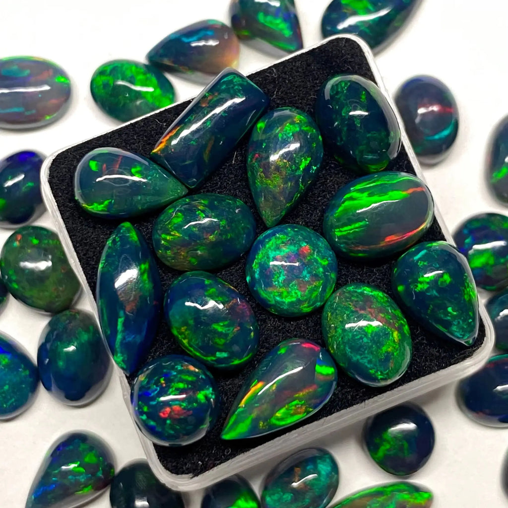 A group of polished teardrop-shaped opal gemstones with vibrant green, blue, and orange flashes, arranged inside a black foam-lined square box with more opals scattered around on a white background.