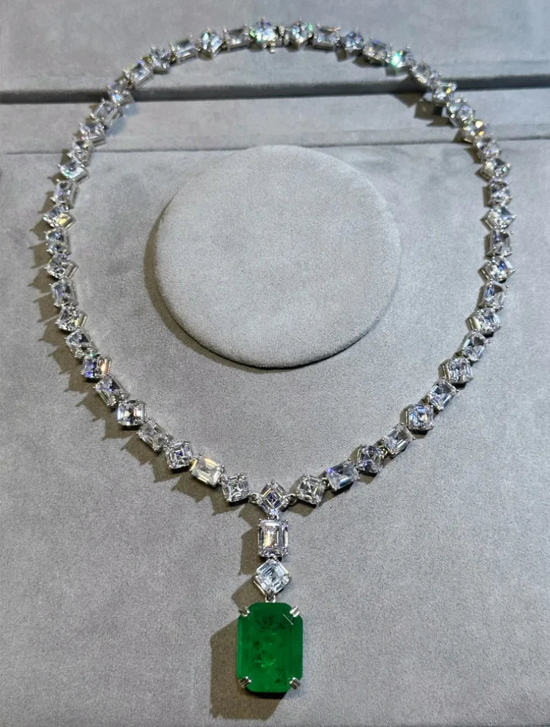 A silver-toned diamond necklace displayed on a gray velvet jewelry stand, featuring a large rectangular emerald-cut green gemstone pendant.