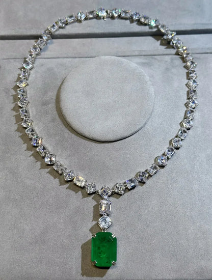 A silver-toned diamond necklace displayed on a gray velvet jewelry stand, featuring a large rectangular emerald-cut green gemstone pendant.
