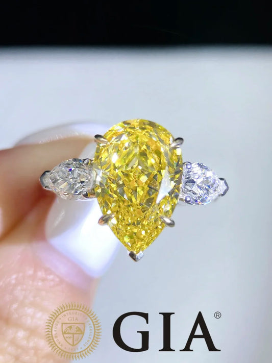 A pear-cut yellow diamond ring held between fingers, featuring two small clear accent diamonds on either side, with a GIA logo visible at the bottom.