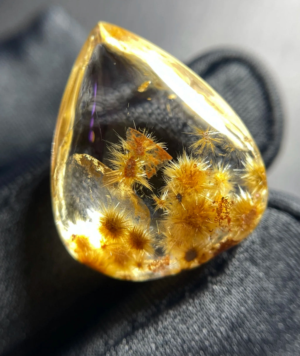 A teardrop-shaped clear gemstone or resin cabochon with golden-yellow crystalline inclusions, photographed close up against a dark gray fabric background.