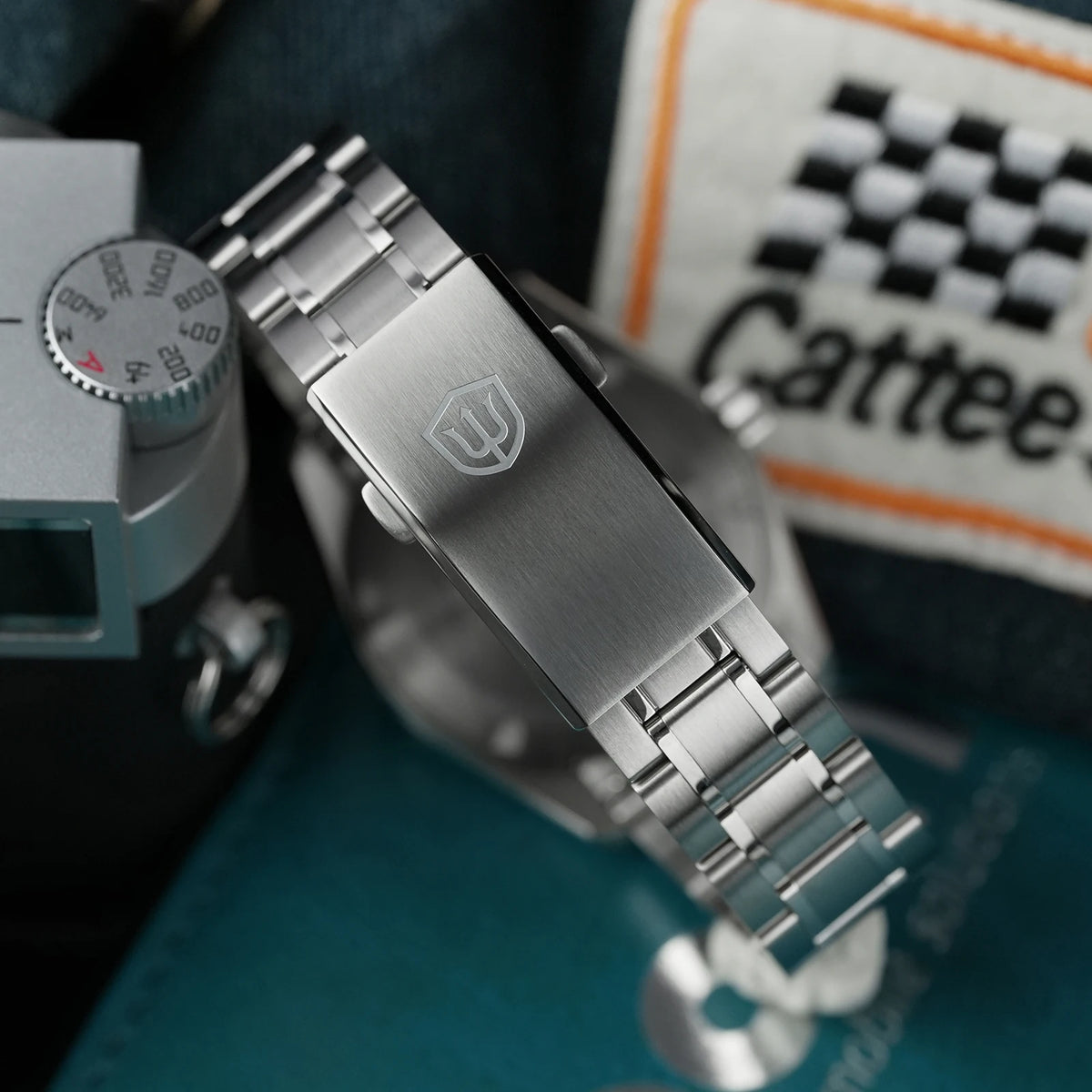A close-up of a stainless steel watch band with a brushed metal clasp and a shield logo, styled on a dark textured surface with a vintage camera and a Cattee patch in the background.