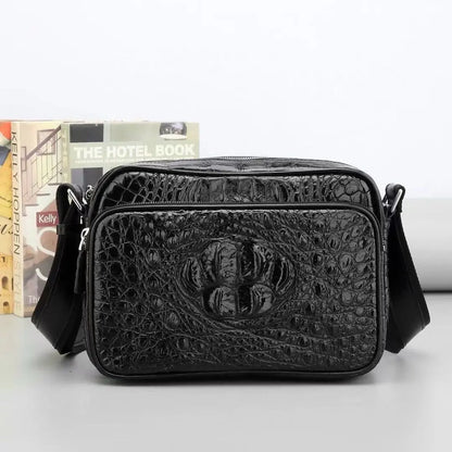 A black crocodile-embossed leather crossbody bag with a front zippered pocket and a central embossed logo, displayed on a light gray surface with books in the background.