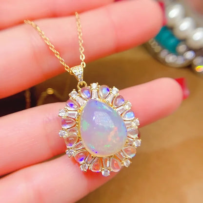 A gold pendant necklace with a teardrop-shaped iridescent opal stone and surrounding clear crystals, held in a person's hand.