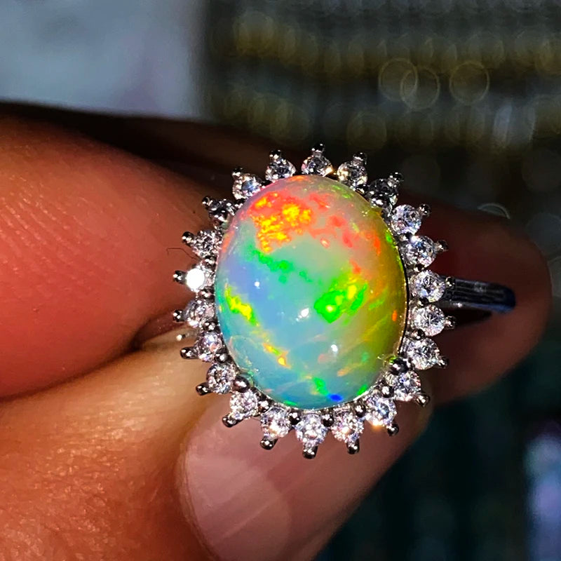 A close-up of a ring held on fingers, featuring a round iridescent opal center stone surrounded by a halo of small clear stones on a silver-toned band.
