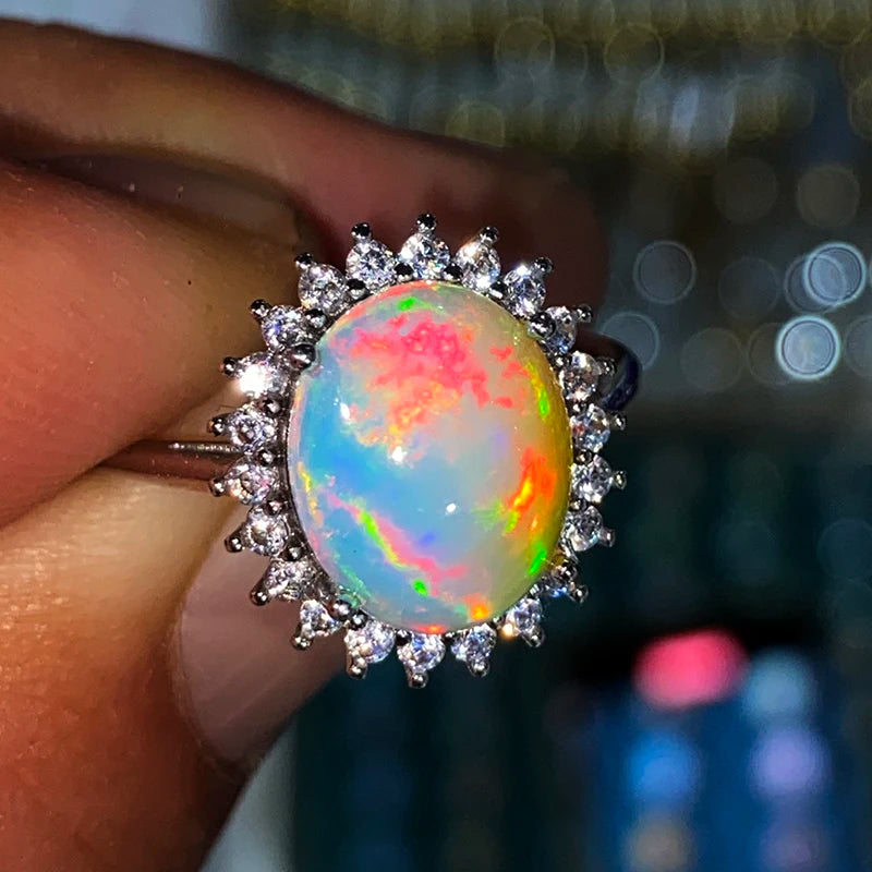 A close-up of a hand holding a silver halo ring featuring a vibrant, color-shifting opal cabochon with blue, pink, green, and orange flashes, surrounded by small clear stones.