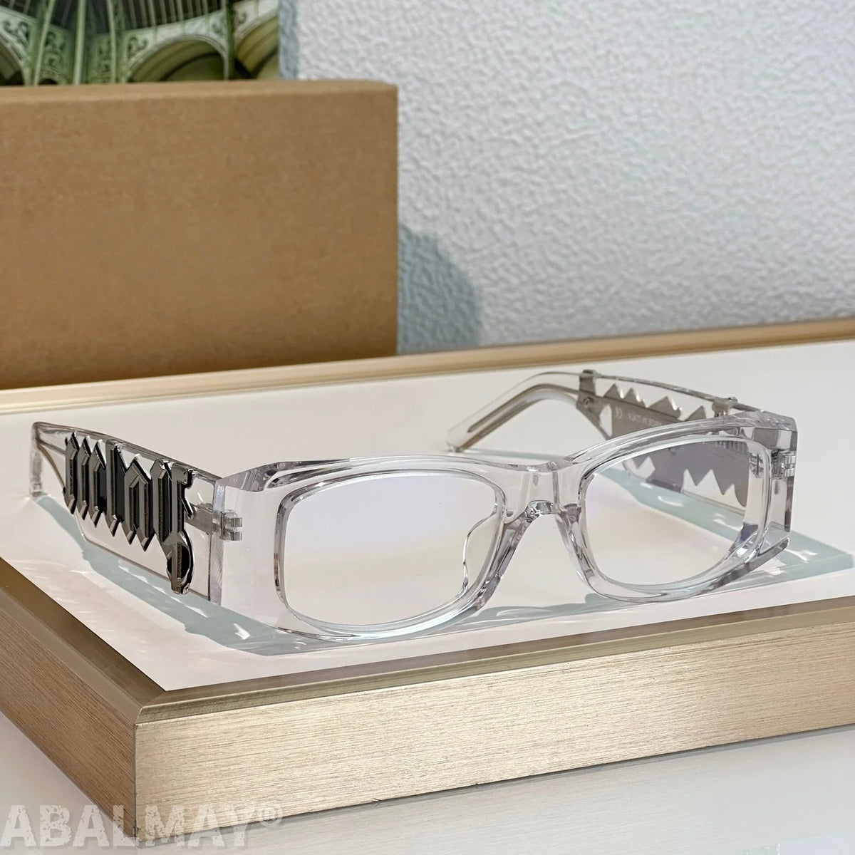 Clear acetate eyeglasses with shark-tooth patterned metal temples, displayed on a gold book with a cardboard box in the background.