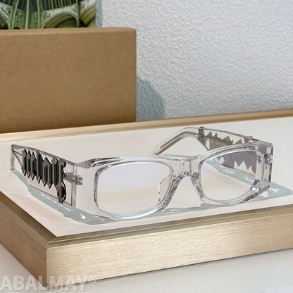 Clear acetate eyeglasses with shark-tooth patterned metal temples, displayed on a gold book with a cardboard box in the background.
