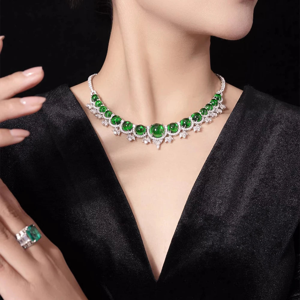 A close-up of a model wearing a silver-toned choker necklace with large emerald-cut green gemstones and small sparkling clear accents, paired with a matching green gemstone ring on a black satin dress against a dark background.