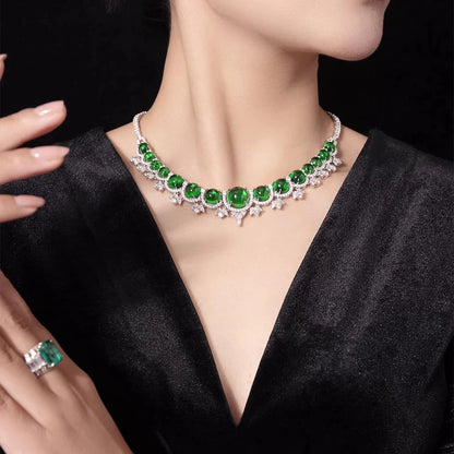 A close-up of a model wearing a silver-toned choker necklace with large emerald-cut green gemstones and small sparkling clear accents, paired with a matching green gemstone ring on a black satin dress against a dark background.