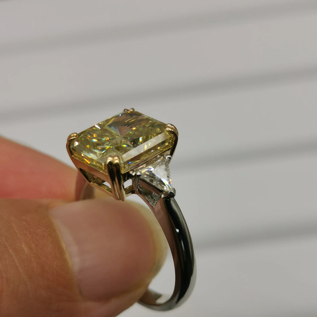 A yellow diamond ring held between fingers. The ring features a large cushion-cut yellow diamond set in a gold-toned four-prong setting, with a smaller clear accent stone on the shank, photographed against a soft-focus gray background.