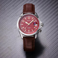 A wristwatch with a silver-toned metal case, a red dial, and a brown leather strap, photographed on a textured gray background.