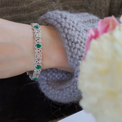 A close-up of a silver-toned bracelet with green and clear gemstones worn on a wrist, styled with a gray knit sleeve and a blurred pink and white flower in the foreground.
