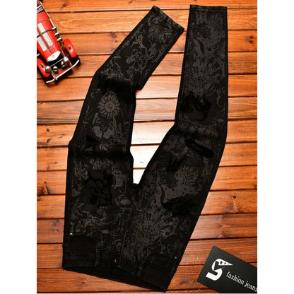 Black denim jeans with a dark gray floral and dragon pattern, laid flat on a wooden plank background next to a toy car and a brand tag.
