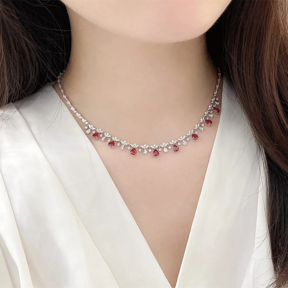 A close-up of a woman wearing a delicate silver-toned necklace adorned with clear crystals and small red gemstone accents, paired with a white satin blouse.