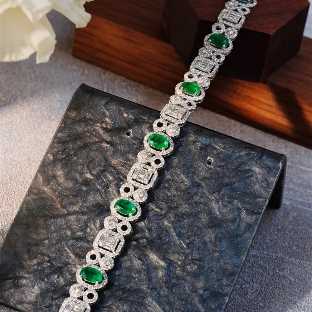 A silver-toned bracelet with oval green emerald-cut stones and round clear stones in a halo setting, displayed on a dark textured stand.