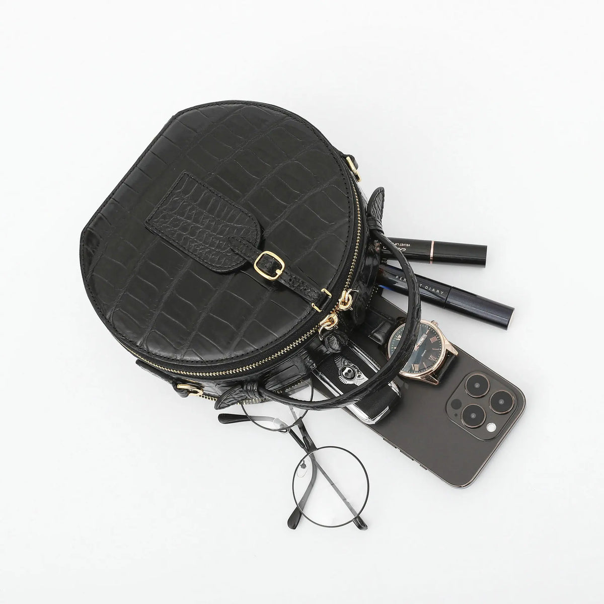 A black crocodile-embossed zippered pouch bag with gold hardware, styled with various everyday accessories including a smartphone, eyeglasses, a watch, and pens on a clean white background.