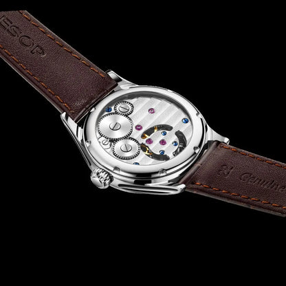 A close-up of a wristwatch with a brown leather strap and a silver-toned case showing the internal mechanical movement through a transparent back, set against a solid black background.