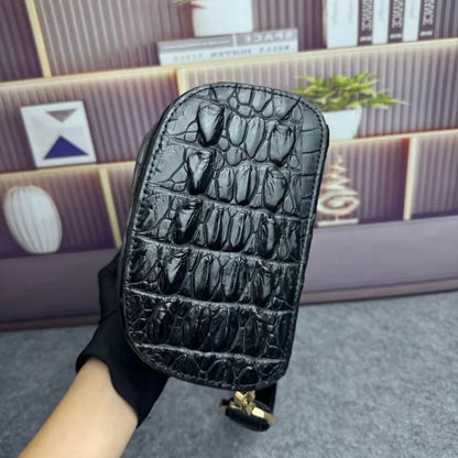 A black crocodile-embossed leather handbag held by a hand wearing a black glove, set against a blurred background of a wooden shelf and books.