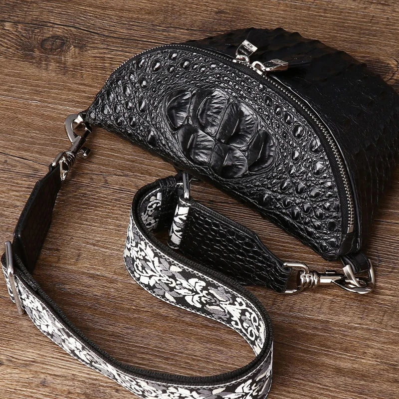Black crocodile-embossed leather crossbody bag with a silver floral-patterned detachable shoulder strap, displayed on a rustic wooden background.