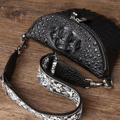 Black crocodile-embossed leather crossbody bag with a silver floral-patterned detachable shoulder strap, displayed on a rustic wooden background.