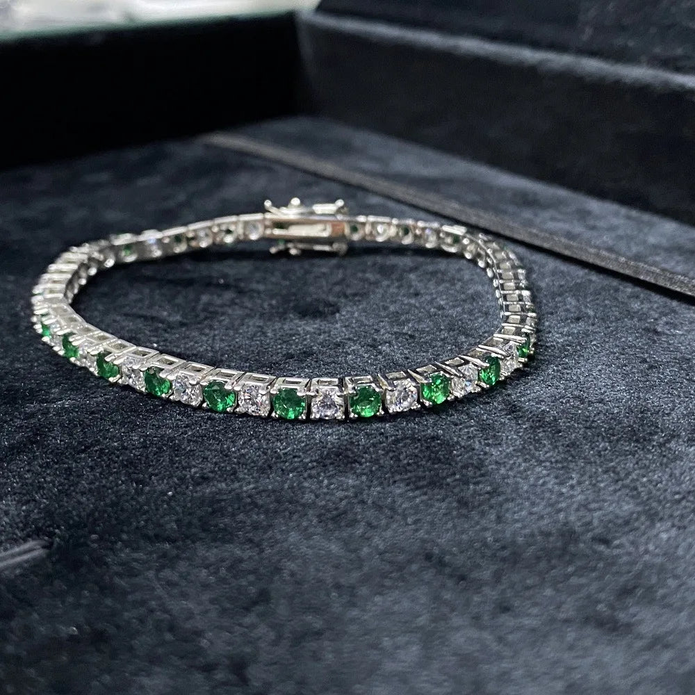 A tennis bracelet with square-cut green emeralds and clear stones set in silver-toned metal, displayed on a black velvet jewelry box.